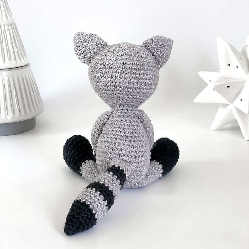 Puede incluir: Un juguete de peluche de mapache de crochet gris con una cola rayada de negro y gris. El juguete est&aacute; sentado sobre una superficie blanca.