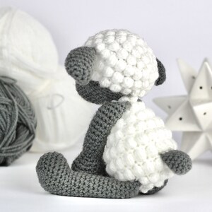 May include: A crocheted white and gray sheep toy with a fluffy white body and gray legs and head. The sheep is sitting on a white surface.