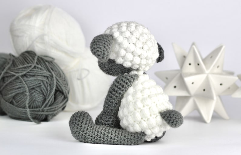 May include: A crocheted white and gray sheep toy with a fluffy white body and gray legs and head. The sheep is sitting on a white surface.