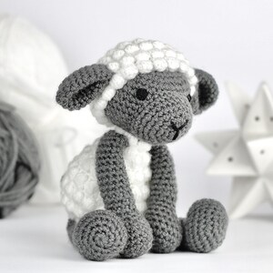 May include: A crocheted gray and white sheep toy with a white fluffy head and gray body. The sheep is sitting on a white surface.
