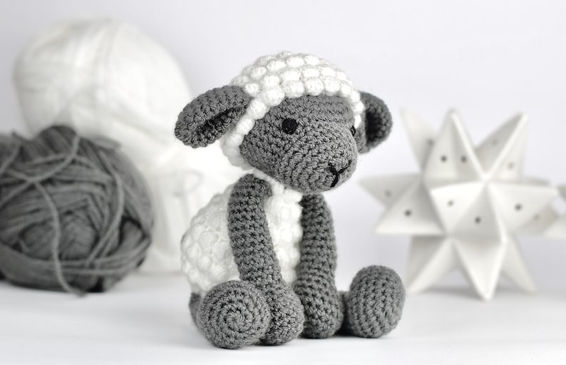 May include: A crocheted gray and white sheep toy with a white fluffy head and gray body. The sheep is sitting on a white surface.
