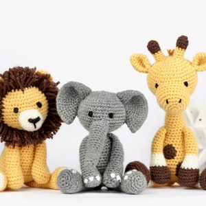 May include: Three crocheted stuffed animals: a lion with a brown mane, a gray elephant, and a yellow giraffe. The animals are sitting on a white surface.