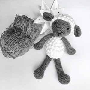 May include: A crocheted gray and white sheep toy with a white face and black eyes. The sheep is sitting on a white surface.