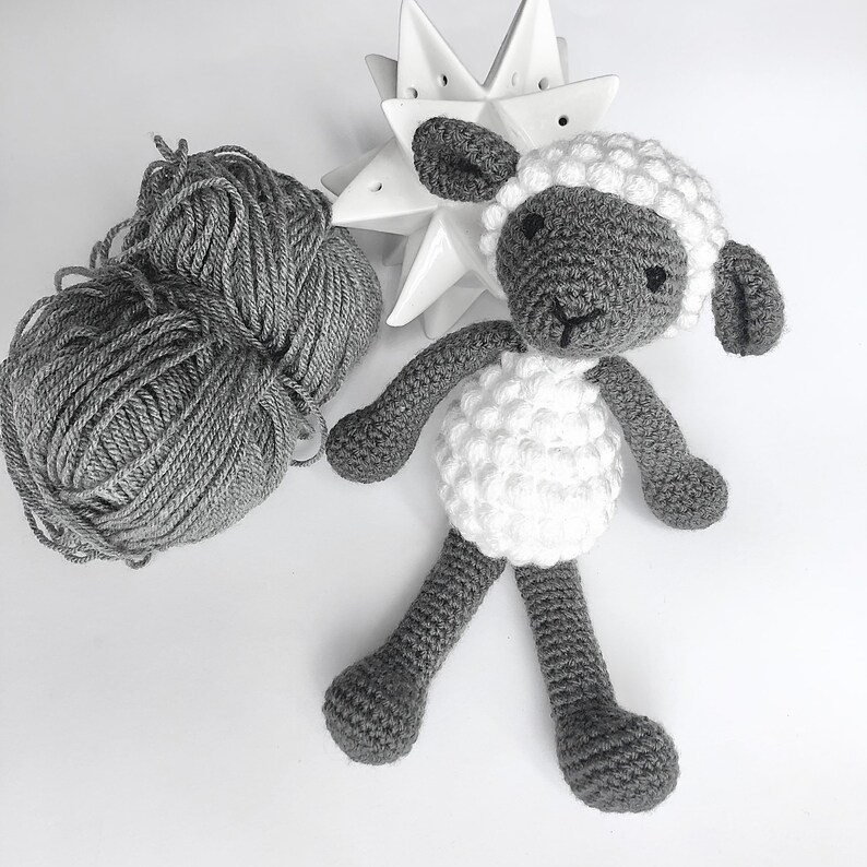 May include: A crocheted gray and white sheep toy with a white face and black eyes. The sheep is sitting on a white surface.