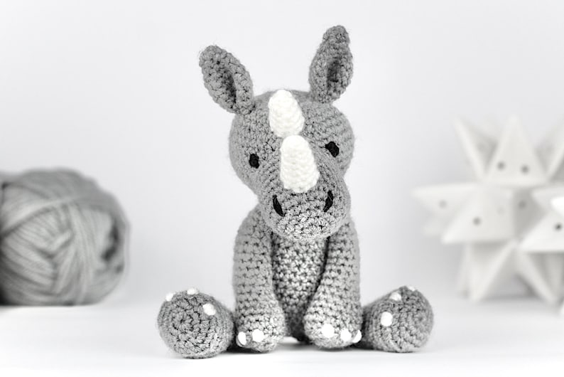 Pode incluir: Um brinquedo de rinoceronte de croch&ecirc; cinza com detalhes brancos. O brinquedo est&aacute; sentado em uma superf&iacute;cie branca.