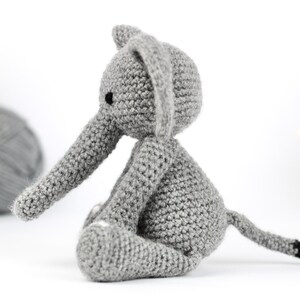 May include: A gray crocheted elephant sitting on a white surface. The elephant has a long trunk, small ears, and a tail with a black tip.