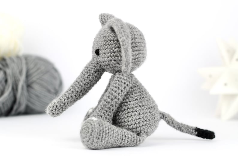 May include: A gray crocheted elephant sitting on a white surface. The elephant has a long trunk, small ears, and a tail with a black tip.