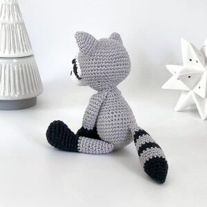 Puede incluir: Un juguete de mapache de crochet con un cuerpo gris, una cola rayada de blanco y negro y patas negras. El juguete est&aacute; sentado sobre una superficie blanca.