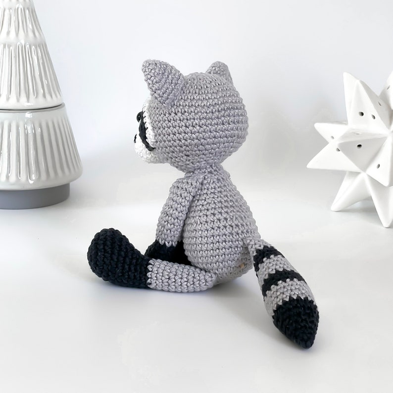 Puede incluir: Un juguete de mapache de crochet con un cuerpo gris, una cola rayada de blanco y negro y patas negras. El juguete est&aacute; sentado sobre una superficie blanca.