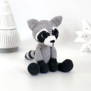 Puede incluir: Un juguete de mapache de crochet con marcas faciales blancas y negras, pelaje gris y patas negras. El juguete est&aacute; sentado sobre una superficie blanca.