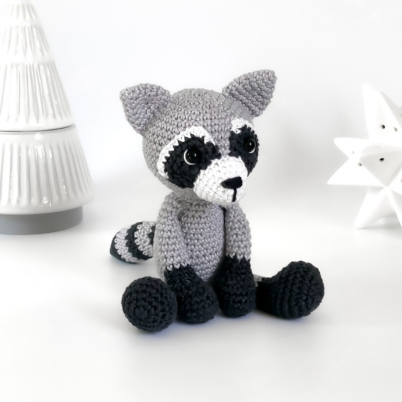Puede incluir: Un juguete de mapache de crochet con marcas faciales blancas y negras, pelaje gris y patas negras. El juguete est&aacute; sentado sobre una superficie blanca.