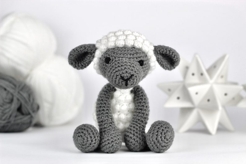 May include: A gray and white crocheted sheep toy with a fluffy white fleece. The sheep has a small black nose and black eyes.
