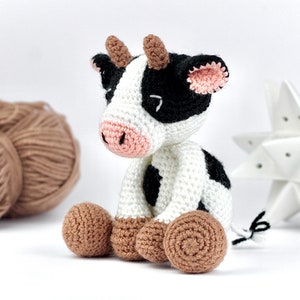May include: A crocheted cow toy featuring a white body with black patches, a pink snout, and brown horns. The cow has brown feet, black ears with pink inner detailing, and a small black and white tail. The toy is crafted with visible yarn stitches.