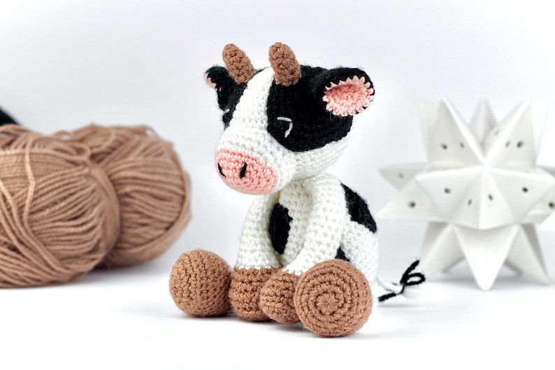 May include: A crocheted cow toy featuring a white body with black patches, a pink snout, and brown horns. The cow has brown feet, black ears with pink inner detailing, and a small black and white tail. The toy is crafted with visible yarn stitches.