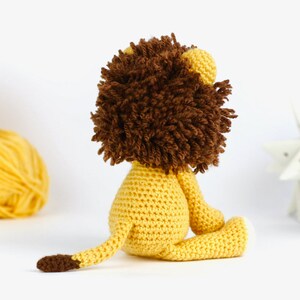 May include: A crocheted lion toy with a brown mane and yellow body. The lion is sitting on a white surface.