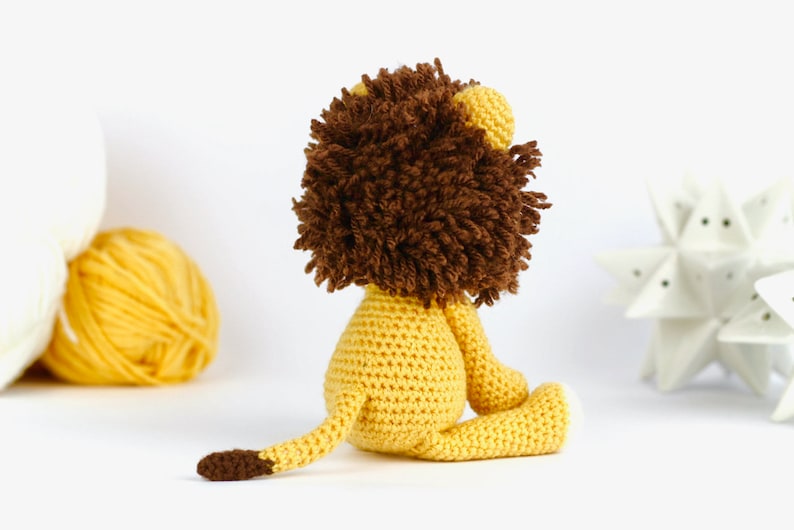 May include: A crocheted lion toy with a brown mane and yellow body. The lion is sitting on a white surface.
