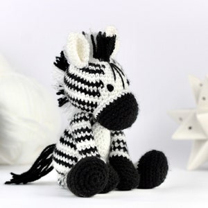 May include: A black and white crocheted zebra soft toy sitting on a white surface. The zebra has a black mane and tail, and black hooves.