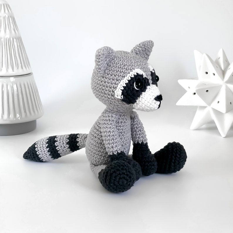 Puede incluir: Un juguete de peluche de mapache tejido a crochet con un cuerpo gris, una cola rayada en blanco y negro y ojos negros. El juguete est&aacute; sentado sobre una superficie blanca.