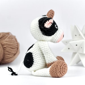 May include: A small, handmade crochet cow toy featuring a white body with black spots, a light brown horn, and light brown hooves. The cow has a pink snout, black ears with pink inner details, and a thin white tail with a small black tuft at the end.