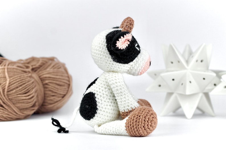 May include: A small, handmade crochet cow toy featuring a white body with black spots, a light brown horn, and light brown hooves. The cow has a pink snout, black ears with pink inner details, and a thin white tail with a small black tuft at the end.