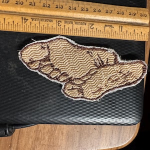 Op de afbeelding: Geborduurde patch in de vorm van een voet, met een bruin en tan chevron patroon. De patch heeft een bruine rand en ligt op een zwart oppervlak. Een houten liniaal met inch-maten staat op de achtergrond.