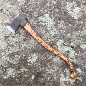 May include: A wooden-handled axe with a dark gray metal head. The axe has a slightly curved handle with a brown and black wood grain pattern.