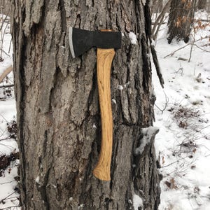 May include: A hand axe featuring a dark, matte-finished metal head with a sharpened silver edge and a curved, light-colored wooden handle with visible grain patterns, positioned vertically against a textured, rugged tree bark surface.