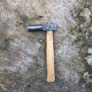 以下が含まれることがあります： 木製の柄が付いた小さな黒い金属製のハンマー。ハンマーは丸い頭とわずかに曲がった柄をしています。