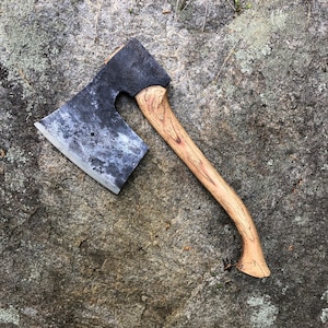 May include: A vintage-style axe with a weathered, dark metal head and a light-colored wooden handle. The axe head has a slightly curved blade and a textured surface. The handle is curved for grip and shows the natural wood grain.