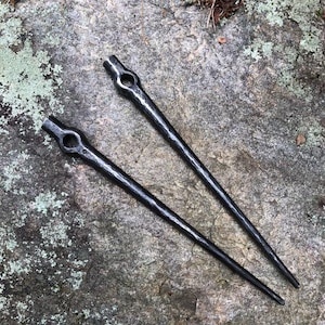 May include: Two hand-forged, dark metal hair sticks. Each stick has a rounded top with a circular opening and a tapered, pointed end. The sticks are displayed on a textured, gray stone surface.