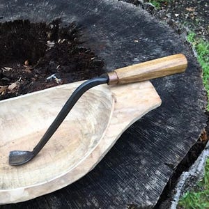 May include: A wooden handled carving tool with a curved metal blade. The tool is resting on a wooden bowl.