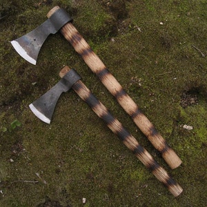 May include: Two hand-forged axes with black handles. The handles are wrapped in a black and brown striped pattern. The axes are on a bed of green moss.