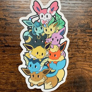 Puede incluir: Una pegatina con una pila de ocho evoluciones de Eevee de dibujos animados. Las evoluciones están ordenadas de mayor a menor, con Sylveon en la parte superior, seguido de Leafeon, Espeon, Umbreon, Jolteon, Flareon, Vaporeon y Eevee.