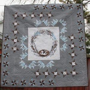 May include: A grey and white quilt with a wreath design in the centre. The wreath is made of twigs and feathers and has a bird's nest in the centre. The quilt is surrounded by a border of grey and white stars and pinwheels.