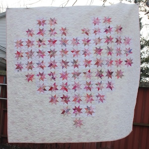 May include: A handmade quilt with a heart design made from pink and red star-shaped patches. The quilt is set against a cream background and has a quilted texture. The quilt is likely to be approximately 180 cm x 180 cm.