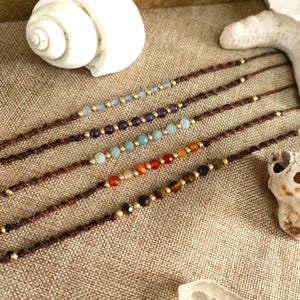 Puede incluir: Primer plano de varias pulseras de cuentas con varias piedras preciosas de colores y cuentas doradas. Las pulseras están ensartadas en cordones marrones. El fondo incluye una concha, un cristal y una superficie de madera.