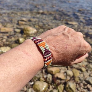 Puede incluir: Pulsera de amistad hecha a mano con un patrón tejido. La pulsera presenta una combinación de colores, incluyendo naranja, burdeos y verde claro. La pulsera se muestra en una muñeca, con un fondo de rocas y agua.
