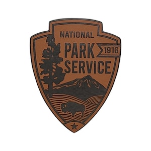 Puede incluir: Un parche de cuero marrón con una silueta negra de una montaña, un árbol y un bisonte. El texto "NATIONAL PARK SERVICE 1916" está impreso en negro.