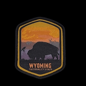 Puede incluir: Un parche bordado en negro y amarillo con una silueta de bisonte frente a una puesta de sol. El texto "WYOMING THE EQUALITY STATE" está impreso debajo del bisonte.