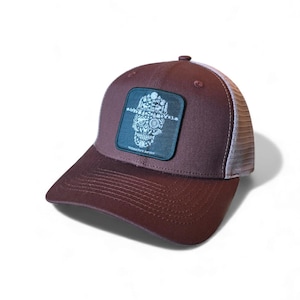 Peut inclure: Casquette de camionneur marron avec un dos en filet blanc et une visière incurvée. Le chapeau présente un écusson carré avec un motif de crâne fait de fleurs et d'engrenages, avec le texte "National Park Service".