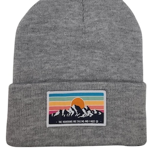 Puede incluir: Gorro de punto gris con un parche de colores con una escena de montaña. El parche presenta una puesta de sol con un sol amarillo y una cita que dice "The mountains are calling and I must go."
