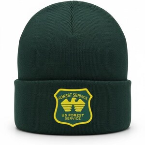 May include: A dark forest green knit beanie with a cuffed brim. Centered on the cuff is a shield-shaped patch with a yellow border, featuring a yellow eagle graphic and the text FOREST SERVICE US FOREST SERVICE in yellow lettering.