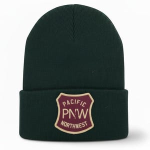 May include: A dark green knit beanie featuring a shield-shaped patch on the cuff. The patch is maroon with a cream border and text that reads PACIFIC PNW NORTHWEST, with the letter N stylized as a pine tree silhouette in cream.