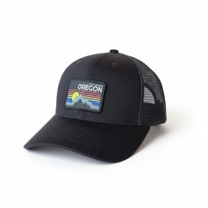 Pode incluir: Boné de caminhoneiro preto com parte traseira em rede e aba curva. O boné apresenta um patch retangular com a palavra "OREGON" acima de um design colorido de montanha e sol. Um estilo clássico, adequado para atividades ao ar livre.