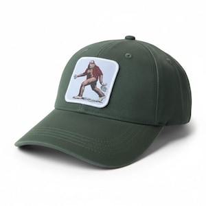 Può includere: Cappellino da baseball verde con visiera curva. Il cappellino presenta una toppa quadrata con un'illustrazione di un Bigfoot con una camicia a quadri e un cappellino da baseball. Realizzato in tessuto resistente.