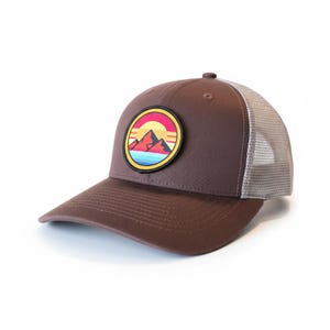 May include: A brown trucker cap with a mesh back and a curved peak. It has a circular patch depicting mountains and a sunset in red, yellow, blue, and pink. A classic style of hat.