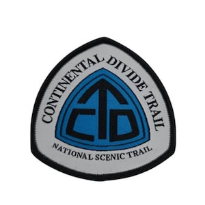 Könnte beinhalten: Weißer Patch mit schwarzem Rand, der ein blaues und schwarzes Logo mit einem nach oben zeigenden Pfeil in einem Dreieck zeigt. Der Text "Continental Divide Trail National Scenic Trail" umgibt das Logo.