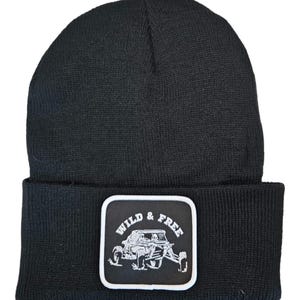 Puede incluir: Gorro de punto negro con un parche blanco que dice "Wild & Free" y presenta una ilustración en blanco y negro de un buggy de dunas.