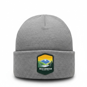Puede incluir: Un gorro de punto gris jaspeado con borde doblado y un parche central. El parche es un hexágono verde oscuro que muestra una ilustración de paisaje con montañas azules, pinos verdes y un cielo amarillo con el texto Rocky Mountain National Park.