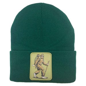 May include: A green knit beanie with a brown and tan patch of a Bigfoot figure carrying a backpack and walking with a stick.